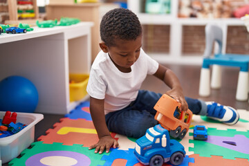 African american boy playing with cars and truck toy sitting on floor at kindergarten