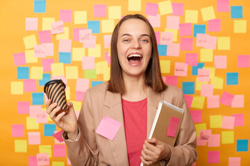 Joyful cheerful exited amazed woman wearing beige jacket posing isolated over yellow background,...