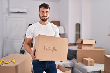 Handsome hispanic man moving to a new home holding fragile box puffing cheeks with funny face. mouth inflated with air, catching air. - obrazy, fototapety, plakaty