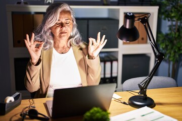 Middle age woman with grey hair working using computer laptop late at night relax and smiling with eyes closed doing meditation gesture with fingers. yoga concept.