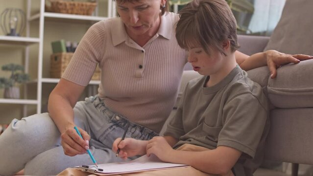 Loving mother caring her daughter with down syndrome with drawing, sitting together on floor in living room