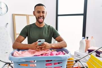 African american man doing laundry using smartphone sticking tongue out happy with funny expression.