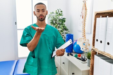 African american physiotherapist man working at pain recovery clinic looking at the camera blowing a kiss with hand on air being lovely and sexy. love expression.
