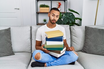 African american young man holding a pile of books sitting on the sofa looking sleepy and tired,...