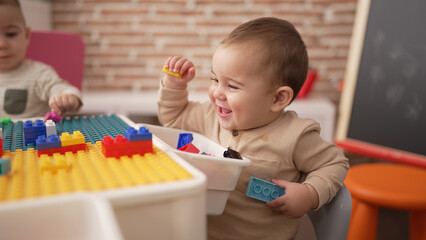Adorable toddler holding plastic construction blocks sitting on table at kindergarten