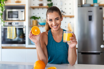 Beautiful young woman drinking fresh orange juice in kitchen. Healthy diet. Happy young woman with glass of juice and orange at table in kitchen. - obrazy, fototapety, plakaty