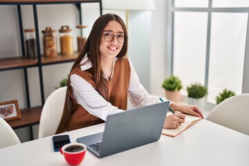 Young beautiful hispanic woman writing on notebook sitting on table studying at home