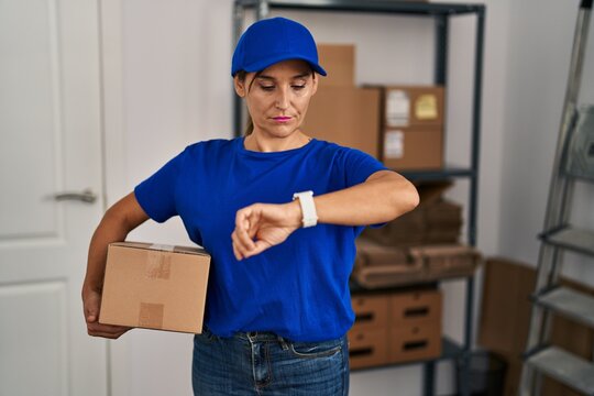 Middle age brunette woman working wearing delivery uniform and cap checking the time on wrist watch, relaxed and confident