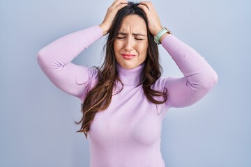 Young brunette woman standing over blue background suffering from headache desperate and stressed because pain and migraine. hands on head.