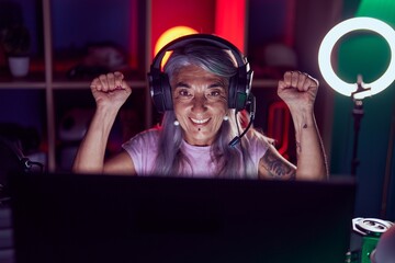Middle age grey-haired woman streamer playing video game with winner expression at gaming room