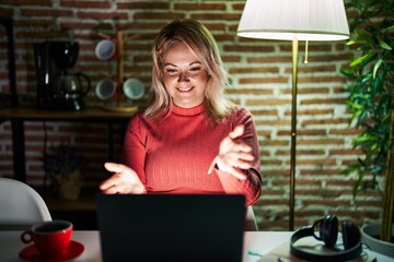 Fototapeta na wymiar Blonde woman using laptop at night at home looking at the camera smiling with open arms for hug. cheerful expression embracing happiness.