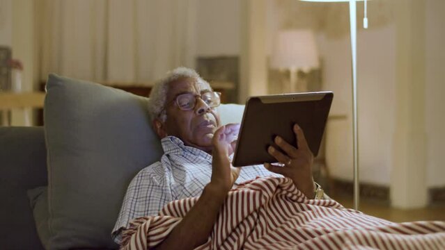 Elderly black man lying in bed surfing net using digital tablet. Grey-haired man in glasses typing on his tablet, web browsing before falling asleep. Leisure, modern technology concept.