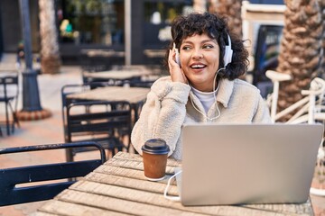 Young beautiful hispanic woman listening to music sitting on table at coffee shop terrace