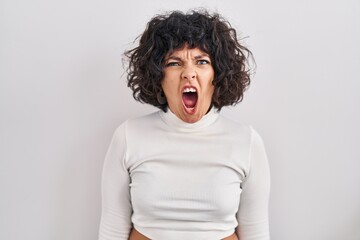 Hispanic woman with curly hair standing over isolated background angry and mad screaming frustrated and furious, shouting with anger. rage and aggressive concept.