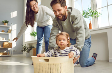 Pastime. Cheerful young parents are playing with their little daughter, carrying her around house in plastic box. Caucasian husband and wife having fun at home on weekend with their toddler child.