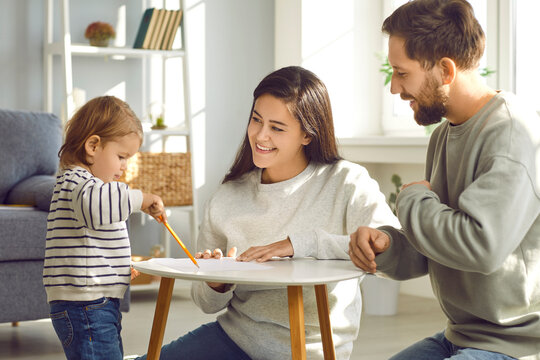 Cute toddler baby drawing on paper with his parents. Cute little girl standing at table with pencil. Happy family spending time together at home, mom and dad helping kid learning to draw and playing