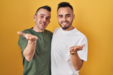 Homosexual couple standing over yellow background smiling cheerful presenting and pointing with palm of hand looking at the camera.