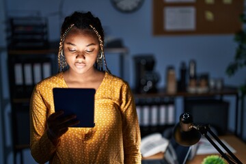 African american woman with braids working at the office at night with tablet relaxed with serious...
