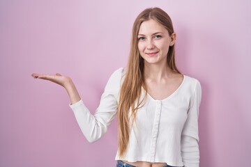 Young caucasian woman standing over pink background smiling cheerful presenting and pointing with palm of hand looking at the camera.