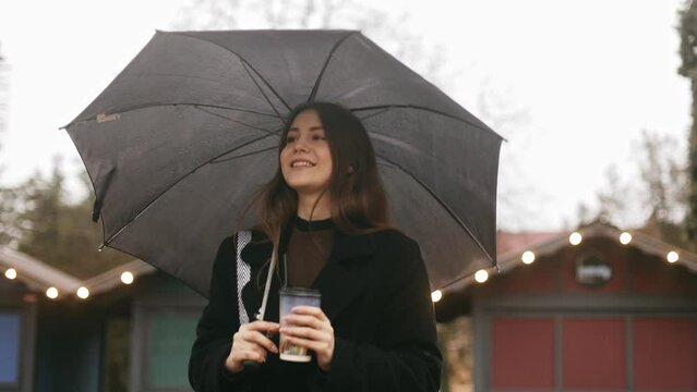 Young cute smiling attractive woman girl goes for a walk on the city street in the rain, holds a black umbrella and a cup of coffee, looks around, loves the rain
