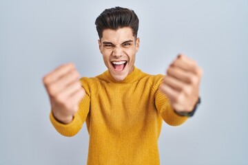 Fototapeta premium Young hispanic man standing over blue background angry and mad raising fists frustrated and furious while shouting with anger. rage and aggressive concept.