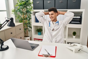 Young hispanic man wearing doctor uniform relaxed with hands on head at clinic