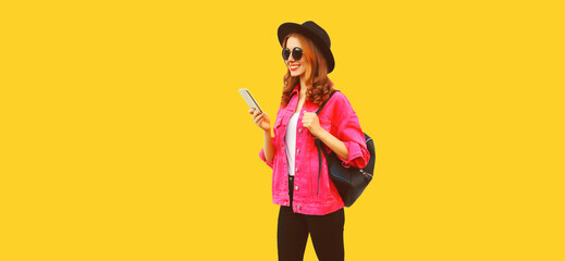 Portrait of stylish modern smiling young woman holding smartphone wearing pink jacket, black round hat on yellow background, blank copy space for advertising text