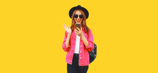 Portrait of stylish modern smiling young woman holding smartphone wearing pink jacket, black round hat on yellow background, blank copy space for advertising text