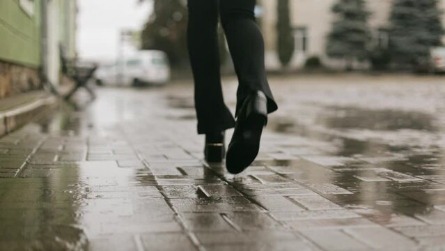 Back view of female slim legs in boots and stylish pants walking on cobblestones on city street in the rain, walking in the rain