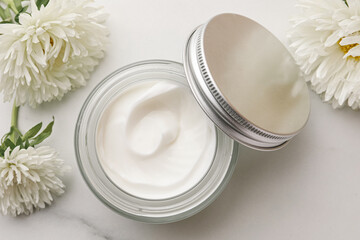 Glass jar of face cream and flowers on white marble table, flat lay