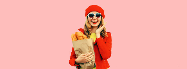 Portrait of happy smiling young woman holding grocery shopping paper bag with long white bread baguette wearing red french beret, heart shaped sunglasses on pink background