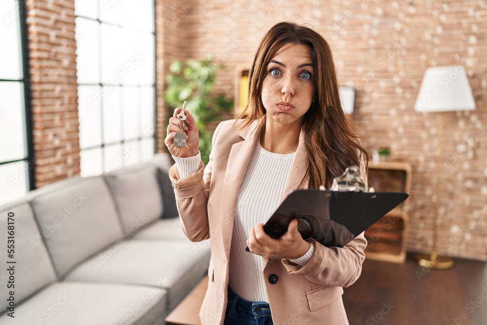 Poster Young brunette woman working as real state agent holding keys of new home puffing cheeks with funny face. mouth inflated with air, catching air.