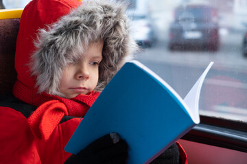 a six-year-old boy rides bus or tram and reads book. Cold season.
