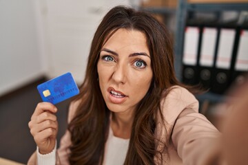 Young brunette woman working at small business ecommerce holding credit card clueless and confused expression. doubt concept.