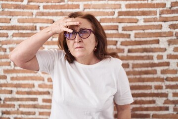 Senior woman with glasses standing over bricks wall pointing unhappy to pimple on forehead, ugly infection of blackhead. acne and skin problem