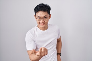 Young asian man standing over white background beckoning come here gesture with hand inviting welcoming happy and smiling