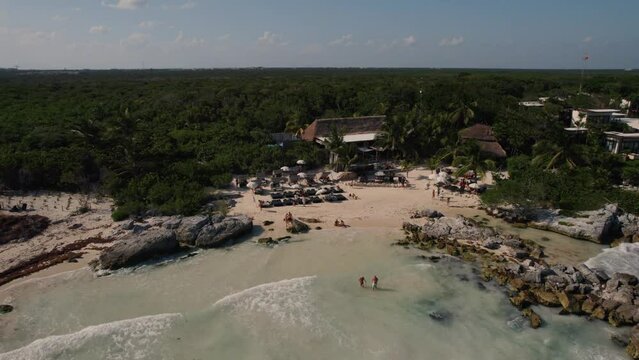 Aerial backwards shot of Beautiful bay with sandy beach and relaxing people during sunny day - Idyllic green landscape island in background - Tulum,Mexico