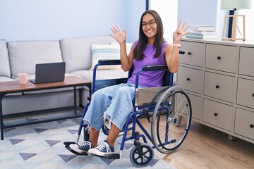 Young hispanic woman sitting on wheelchair at home showing and pointing up with fingers number nine while smiling confident and happy.