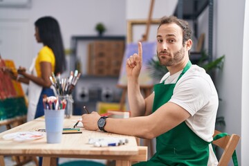 Hispanic man at painter studio smiling with an idea or question pointing finger with happy face, number one