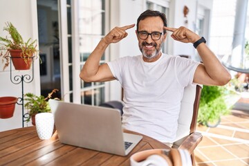 Middle age man using computer laptop at home smiling pointing to head with both hands finger, great idea or thought, good memory