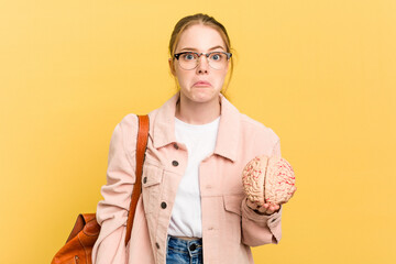 Young student woman holding a brain isolated on yellow background shrugs shoulders and open eyes...