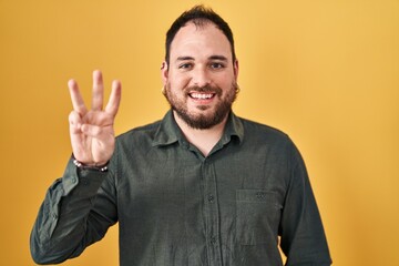 Fototapeta premium Plus size hispanic man with beard standing over yellow background showing and pointing up with fingers number three while smiling confident and happy.