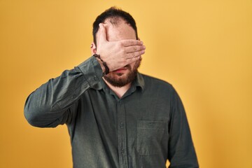 Plus size hispanic man with beard standing over yellow background covering eyes with hand, looking serious and sad. sightless, hiding and rejection concept