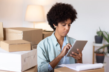 Logistics manager examining parcel tracking. Serious blackwoman with curly hair sitting among stacks of boxes and using tablet. Postal service or shipping concept