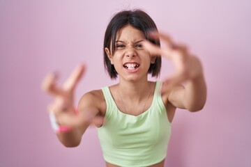 Naklejka premium Young girl standing over pink background shouting frustrated with rage, hands trying to strangle, yelling mad