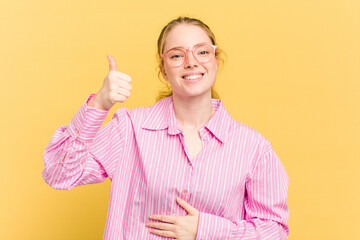 Young caucasian redhead woman isolated on yellow background touches tummy, smiles gently, eating and satisfaction concept.