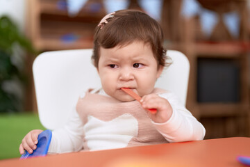 Adorable toddler sitting on table sucking plastic spoon holding dish at kindergarten