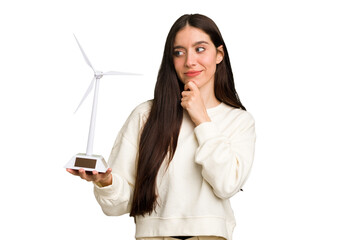 Young caucasian woman holding a small wind energy mill isolated looking sideways with doubtful and...