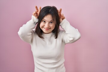 Woman with down syndrome standing over pink background posing funny and crazy with fingers on head as bunny ears, smiling cheerful