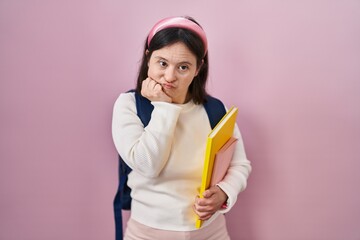 Woman with down syndrome wearing student backpack and holding books looking stressed and nervous with hands on mouth biting nails. anxiety problem.